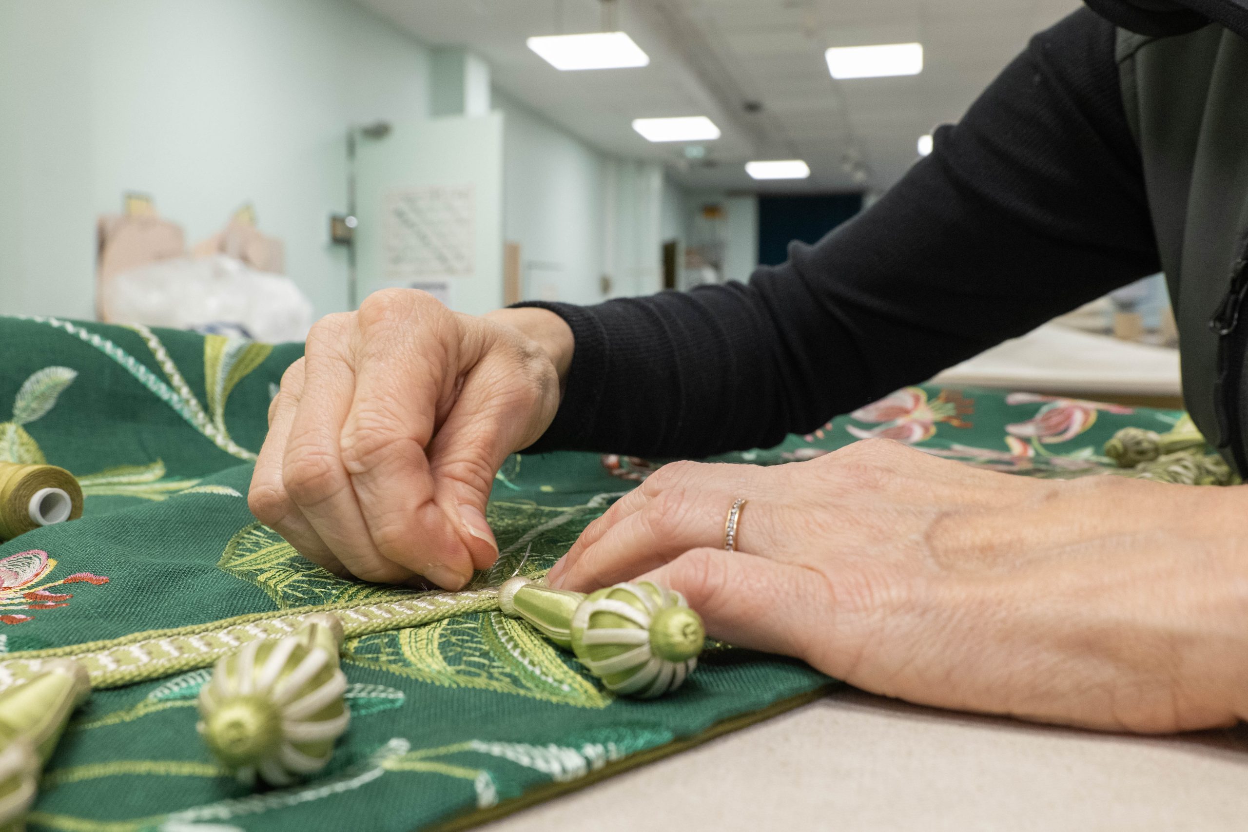 Tapissière posant une passementerie à glands sur un tissu brodé vert à l'atelier